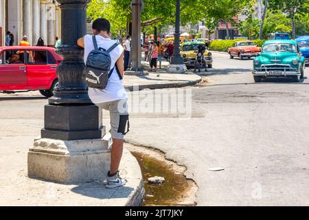 Un giovane si appoggia sul palo di una luce stradale. Un'auto d'epoca guida nel quartiere del centro. L'acqua sporca è stagnante dal marciapiede Foto Stock