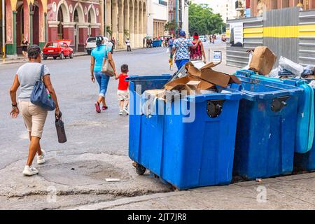 Persone cubane che camminano da grandi bidoni della spazzatura in una strada cittadina. I rifiuti vengono scoperti e traboccanti. Foto Stock