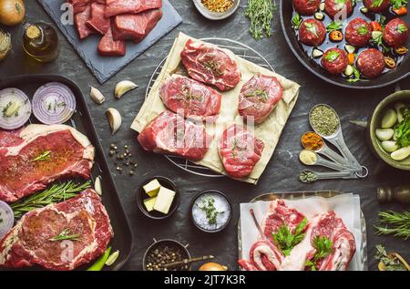 Diversi tipi di carne cruda fresca cosparsi di condimenti e posta con ingredienti biologici su fondo scuro. Vista dall'alto con primo piano. Foto Stock
