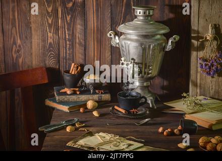 Classica vita morta con samovar russo d'annata posto con una tazza di tè caldo, vecchi libri, noci, bastoncini di cannella e fiori secchi su un tavolo rustico di legno. Foto Stock