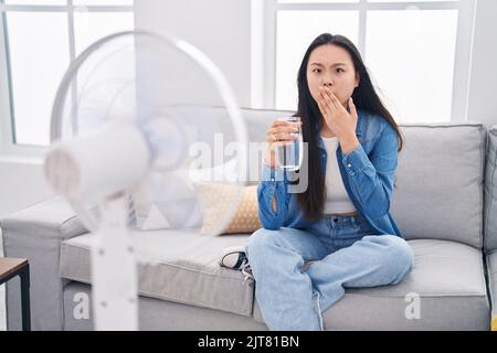 Giovane donna asiatica bevendo un bicchiere d'acqua godendo di aria dal ventilatore che copre la bocca con la mano, scioccato e impaurito per errore. Sorpresa espressione Foto Stock