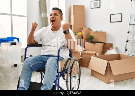 Giovane uomo ispanico seduto su sedia a rotelle a nuova casa molto felice ed eccitato facendo il gesto del vincitore con le braccia sollevate, sorridendo e urlando per il successo. Foto Stock