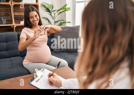 Giovane donna incinta alla sessione di terapia sorridente nell'amore mostrando il simbolo del cuore e la forma con le mani. Concetto romantico. Foto Stock