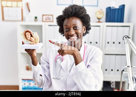 Medico africano donna che tiene modello anatomico di utero femminile con il feto sorridente felice puntamento con mano e dito Foto Stock