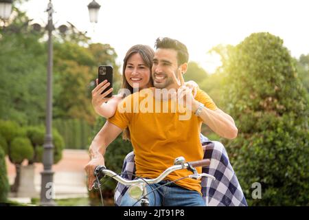 Coppia tenera che prende un selfie mentre cavalca una bici retrò in un parco durante il tramonto Foto Stock