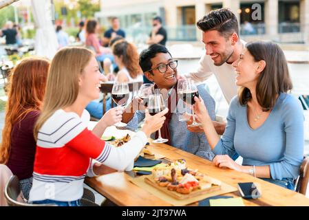 Gruppo di uomini e donne allegri e multirazziali con un bicchiere di vino sorridente e che propone toast seduti al tavolo nel bar all'aperto Foto Stock