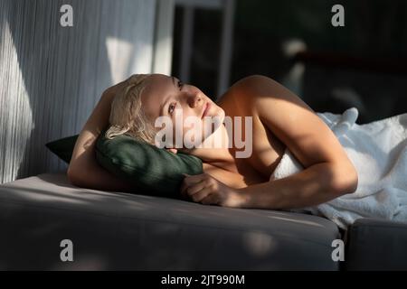 Ritratto di giovane donna piuttosto felice in accappatoio bianco si trova sul letto nella sala spa. Bella ragazza con relax sul letto in luce del giorno Foto Stock