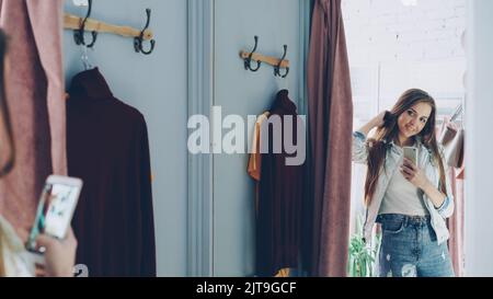 Bella ragazza giovane sta facendo selfie specchio con lo smartphone mentre in piedi in fitting room in negozio di vestiti. Lei sta posando, girando e toccando i suoi lunghi capelli castani con un bel sorriso. Foto Stock