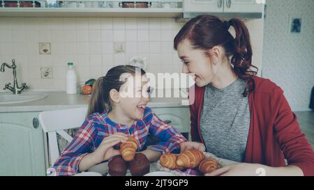 Buona madre e simpatica figlia a colazione mangiando muffin e parlando a casa in cucina moderna. Concetto di famiglia, cibo, casa e persone Foto Stock