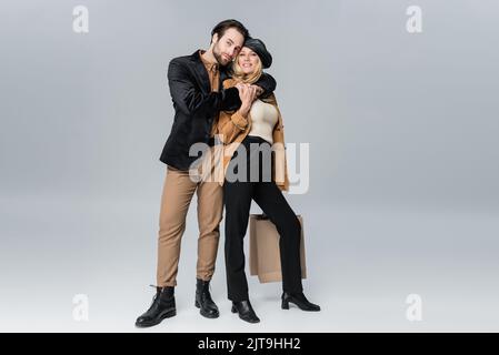 uomo allegro abbracciando donna felice in elegante berretto che tiene borse da shopping sul grigio, immagine stock Foto Stock