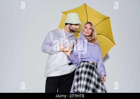 donna in gonna tartan posa con uomo in cappello panama sotto ombrello giallo su grigio, immagine stock Foto Stock