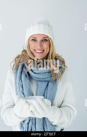 ritratto di donna felice in cappello di inverno e guanti bianchi tenendo una tazza di caffè isolato su grigio, immagine stock Foto Stock