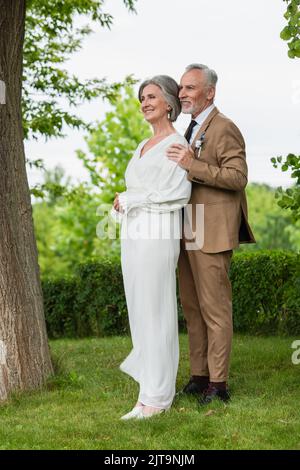 intera lunghezza di sposo felice di mezza età in vestito abbracciando sposa matura in vestito bianco in giardino, immagine stock Foto Stock