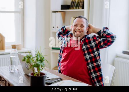 Giovane uomo con sindrome di Down seduto alla scrivania in ufficio e utilizzando un computer portatile, guardando la fotocamera e sorridendo. Foto Stock