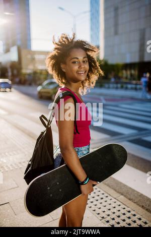 Ragazza adolescente multirazziale con zaino e skateboard, camminando in città durante il giorno estivo. Foto Stock