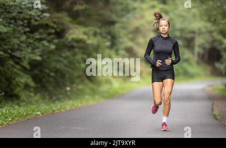 Adatta la giovane ragazza in abbigliamento sportivo nero e scarpe rosse su una strada asfaltata vuota nella natura. Foto Stock