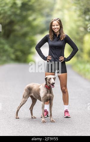 Una giovane donna sportiva e il suo cane Weimaraner stanno insieme nella foresta. Foto Stock