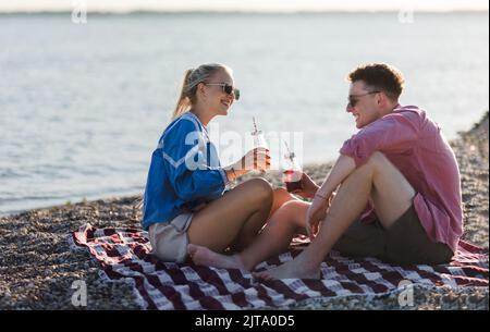 Felice giovane coppia che si riunisce in spiaggia, seduta su una coperta e brindisi con birra in bottiglia. Godersi le vacanze insieme. Foto Stock