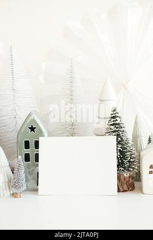 La scheda di Natale mock in su. Biglietto d'auguri vuoto e piccoli alberi di natale e case decorazioni su tavola bianca. Modello cartolina con spazio per il testo e. Foto Stock