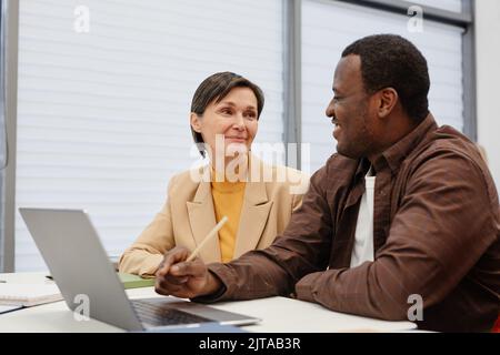 Donna matura che parla con un insegnante africano mentre si siede insieme alla scrivania e imparando un programma informatico sul laptop Foto Stock
