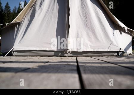 primo piano con vista sul carattere di una tenda glamping, entrata in tessuto bianco su un ponte di legno in un prato verde circondato da una foresta di abeti Foto Stock