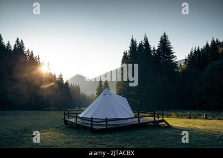 vista posteriore della tenda glamping bianca sul ponte di legno in verde prato circondato da foresta di abeti all'alba Foto Stock