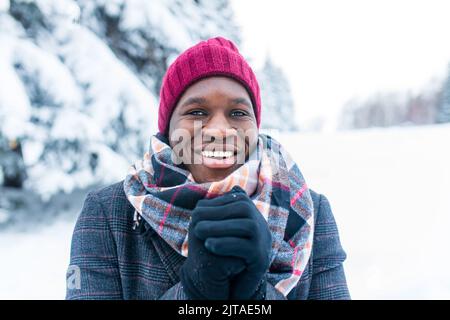 uomo afro-americano in cappello rosso ed elegante cappotto a plaid guardare la macchina fotografica con il sorriso bianco-neve al coperto nel parco Foto Stock