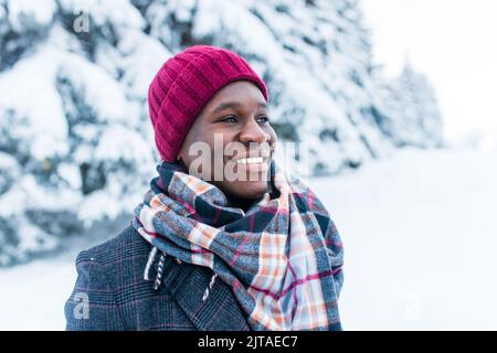 uomo afro-americano in cappello rosso ed elegante cappotto a plaid guardare la macchina fotografica con il sorriso bianco-neve al coperto nel parco Foto Stock