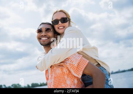 gioioso afroamericano uomo piggybacking donna sorridente e guardando la macchina fotografica Foto Stock