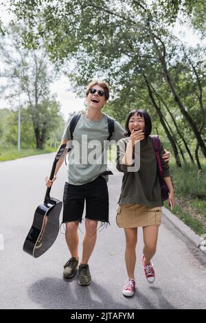 coppia interrazziale eccitata con zaini e chitarra ridere mentre camminano sulla strada Foto Stock