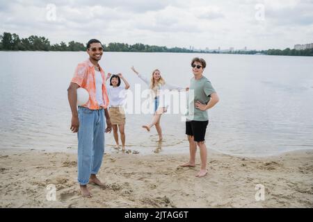 uomo afroamericano che tiene la palla e sorridente alla macchina fotografica vicino agli amici interracial sulla spiaggia Foto Stock