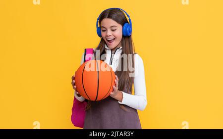 bambino stupito in cuffie con zaino da scuola e palla da basket su sfondo giallo Foto Stock