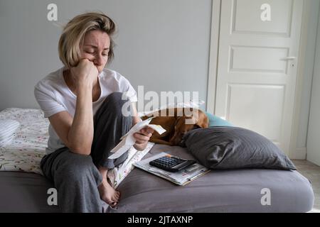 La donna bionda di mezza età rovesciato in pajama si siede sul letto che tiene le fatture non pagate closeup Foto Stock