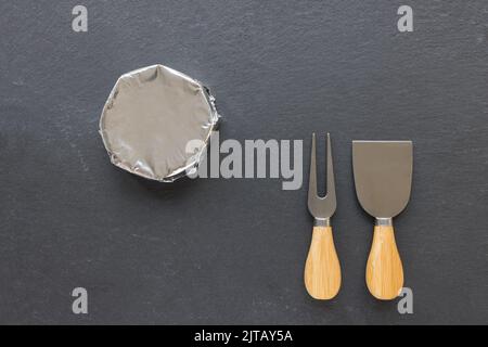 Formaggio francese rotondo camembert in carta stagnola e coltelli formaggio su piatto nero ardesia. Primo piano, vista dall'alto, spazio di copia Foto Stock