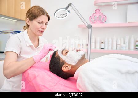Estetista esperto che applica la maschera alla pelle facciale del giovane uomo Foto Stock
