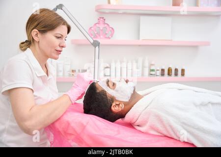 Cosmetician applicare la maschera di caolino alla pelle facciale dell'uomo Foto Stock
