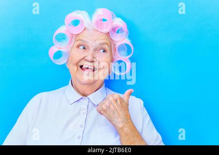 Affascinante vecchia donna con ricciolatori su bianco grigio capelli indossare camicia prepararsi per data isolato su sfondo blu brigth Foto Stock