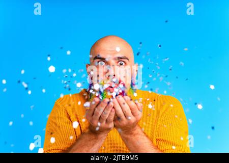 bell'uomo con raindow in barba e maglione giallo che soffia i dolciumi in baclground blu studio Foto Stock