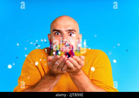 bell'uomo con raindow in barba e maglione giallo che soffia i dolciumi in baclground blu studio Foto Stock