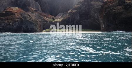 Scogliere erodenti e una piccola spiaggia sabbiosa sulla costa di Napali in contrasto con le acque blu profonde dell'Oceano Pacifico, visto da una barca, in Foto Stock