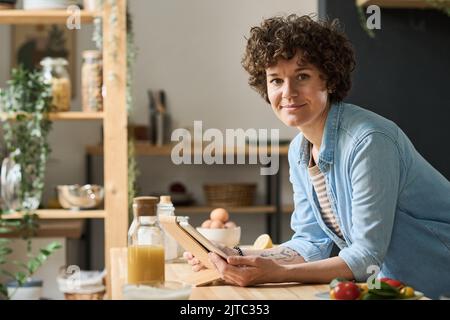Ritratto di una giovane donna con capelli ricci guardando la fotocamera mentre si utilizza un tablet digitale durante la preparazione del cibo Foto Stock