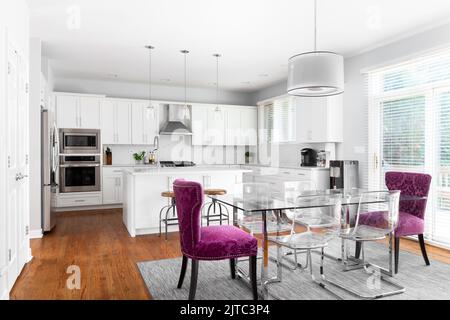 Una lussuosa sala da pranzo e cucina con un tavolo in vetro, sedie trasparenti e viola, armadi bianchi ed elettrodomestici in acciaio inox. Foto Stock