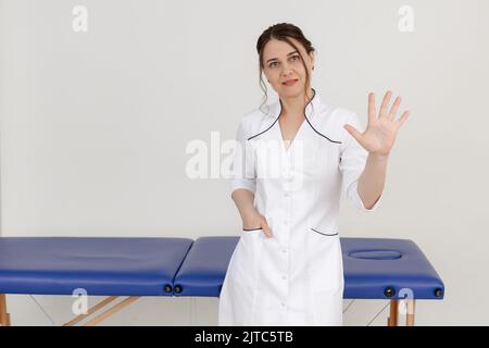 Ritratto di donna massaggiatrice sicura di mezza età in piedi vicino al divano blu, allungando la mano in avanti con palmo aperto. Foto Stock