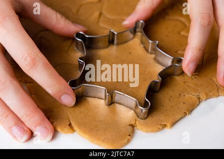 Primo piano mani donna taglio biscotti con muffa metallica dalla pasta frolla. Un metallo si forma sotto forma di fiocchi di neve per fare il pan di zenzero per Natale. Foto Stock
