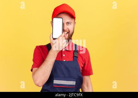 Ritratto di felice sorridente gioioso lavoratore barbuto uomo in piedi e occhio di copertura con telefono cellulare con schermo vuoto per il testo promozionale. Studio al coperto isolato su sfondo giallo. Foto Stock