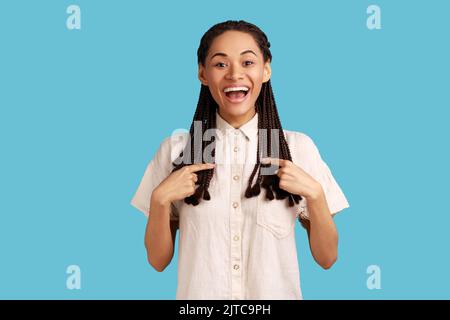 Donna allegra eccitata con greggi neri che puntano a se stessa e sorridendo in generale, estremamente felice di essere scelto, indossando una camicia bianca. Studio in interni isolato su sfondo blu. Foto Stock