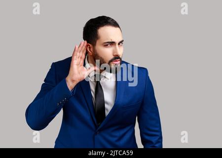 Uomo bearded concentrato nosy che tiene la mano vicino all'orecchio, ascoltando i discorsi interessanti e segreti privati, spionaggio, indossando il vestito ufficiale di stile. Studio in interni isolato su sfondo grigio. Foto Stock