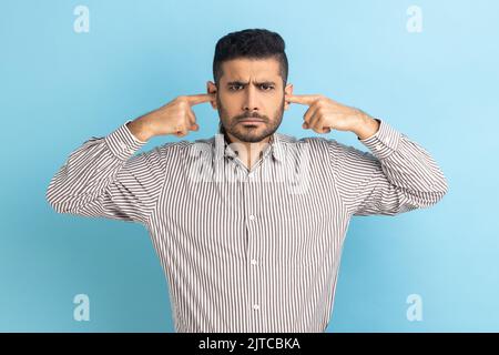 Non voglio sentire questo. Annoiato frustrato businessman chiudere le orecchie con le dita ignorando le informazioni, rumore irritante, indossare camicia a righe. Studio in interni isolato su sfondo blu. Foto Stock