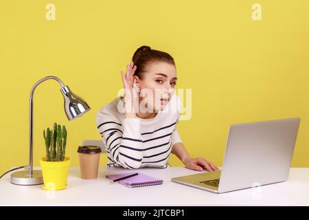 Non ti sento. Donna manager che tiene il braccio vicino all'orecchio cercando di ascoltare il segreto mentre lavora online sul notebook in ufficio. Studio interno girato isolato su sfondo giallo. Foto Stock