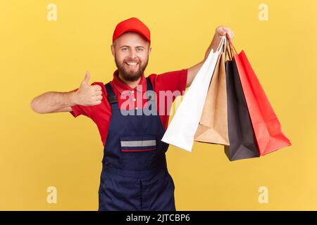 Ritratto di felice sorridente corriere uomo in uniforme tenendo borse della spesa in mano, mostrando pollice in su, consegna rapida dal negozio, industria di servizi. Studio al coperto isolato su sfondo giallo. Foto Stock